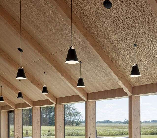 Full indoor shot of a wooden showroom. The ceiling is a light beige, with visible wooden beams angled to create a pitched roof structure.  The ceiling planks have a subtle textured pattern. Seven 'Above' black cone-shaped pendant lights hang from the ceiling. Large windows stretch along the length of the porch, extending from one wall to the other. Through the windows, a wide agricultural landscape of fields is visible. The fields are golden-green. The landscape view stretches from the middle of the image to the horizon in the distance. 