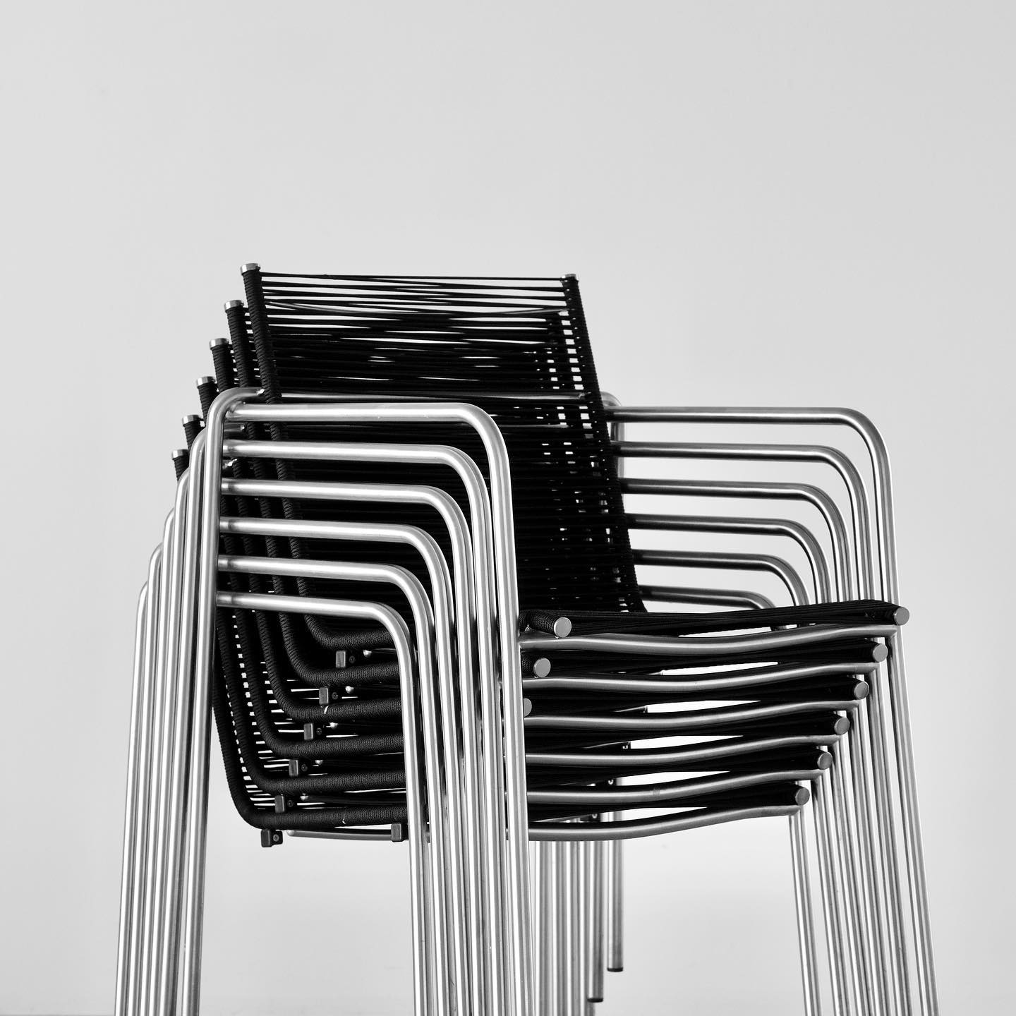 A stack of six chairs, one on top of the other, are shown standing upright. All of the chairs are the same with black woven seats, and chrome steel frames. Only the chair on top is fully visible. The chairs are against a white wall.