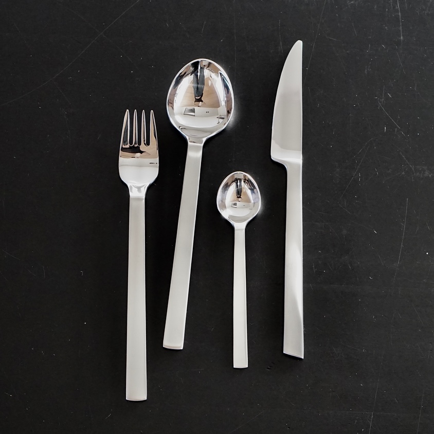 The image shows a fork, knife and two spoons arranged vertically on a black background. The fork, knife and spoons are all made out of silver, polished and shiny. The background is textured with scratches and a subtle sheen of light.