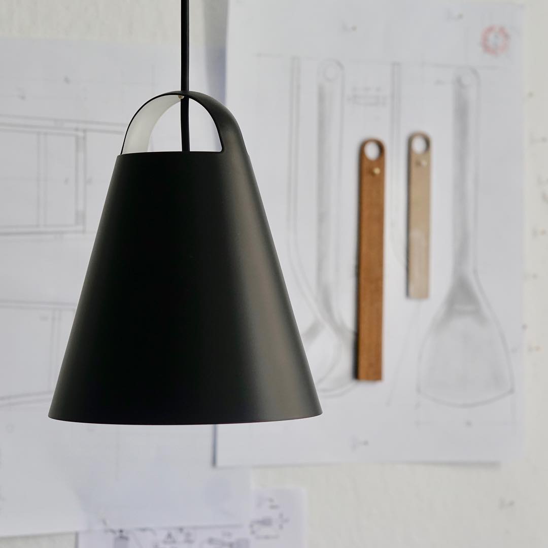 Close-up of a 'Above' lampshade hanging against a white wall. The light fixture's shade is a matte black, featuring a slightly concave, cone-shaped form.  A handle-like cutout is visible near the top of the shade. The wall behind the light fixture is covered with architectural or design drawings, showing various sketches in pencil. Several sketches of objects, seemingly utensils or other items, are visible. There are also a couple of light-brown wooden measuring tools/straightedges in the drawings.  The drawings are slightly out of focus, emphasizing the light fixture.