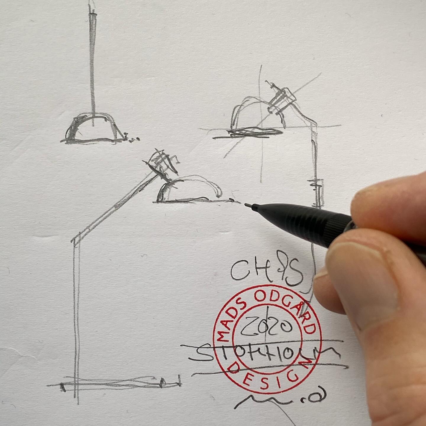 The image shows a hand holding a pen over a sketch of a lamp. The sketch is on a white piece of paper and is done in pencil. The lamp is drawn in a simple fashion, with a base and a shade. Mads Odgård's hand is holding the pen just above the sketch, as if ready to add more details. Below his hand is a red stamp reading 'Mads Odgård Design' and a signature written on top of it.