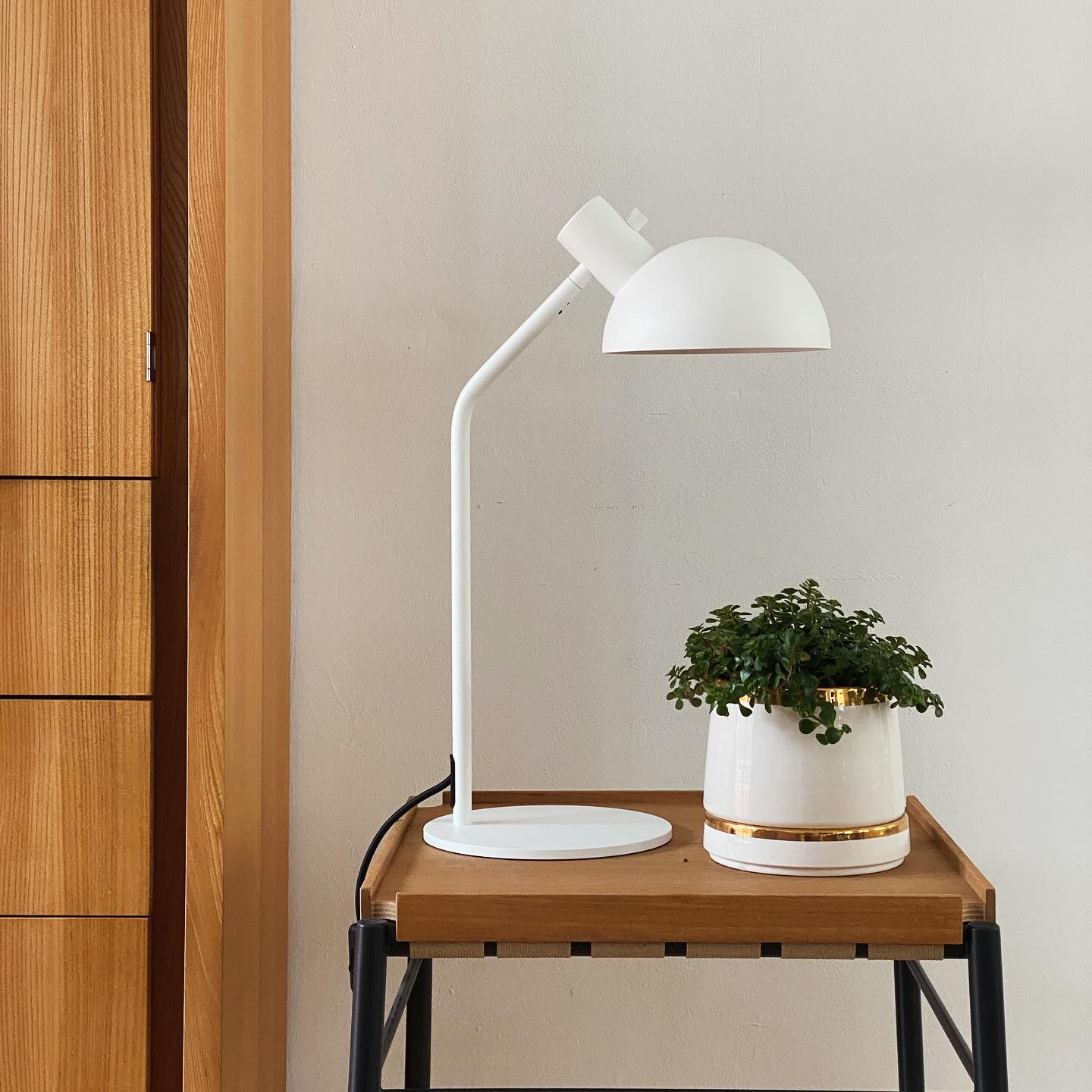 The image shows a white desk lamp on a wooden side table. The lamp is a simple, minimalist design with a white shade and a white base. The lamp is on a wooden side table with a black metal frame that sits on a light colored wood floor. There is also a white pot with a gold rim that has a small green plant in it. The pot is sitting on the table beside a white book. The background of the image is a light-colored wall.