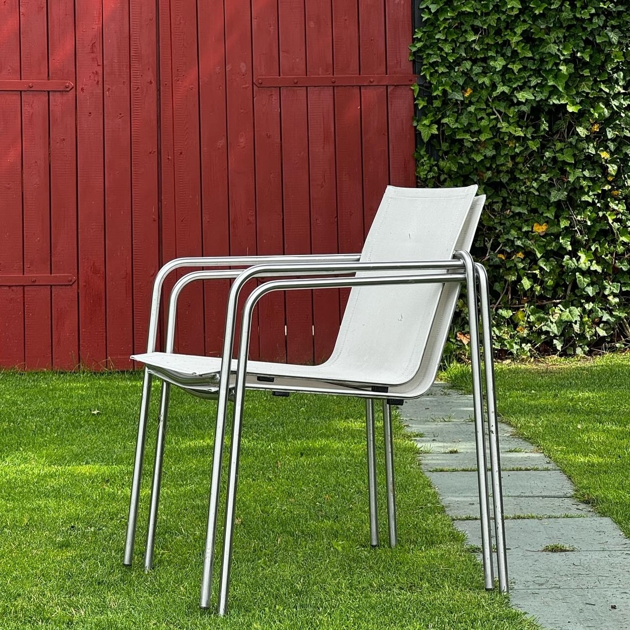 A 'Taku' white metal chair with a white canvas seat sits in the grass. It is positioned to the infront of a red barn and a patch of ivy. On the floor behind the chair, there is a brick walkway lined with large, rectangular stones.