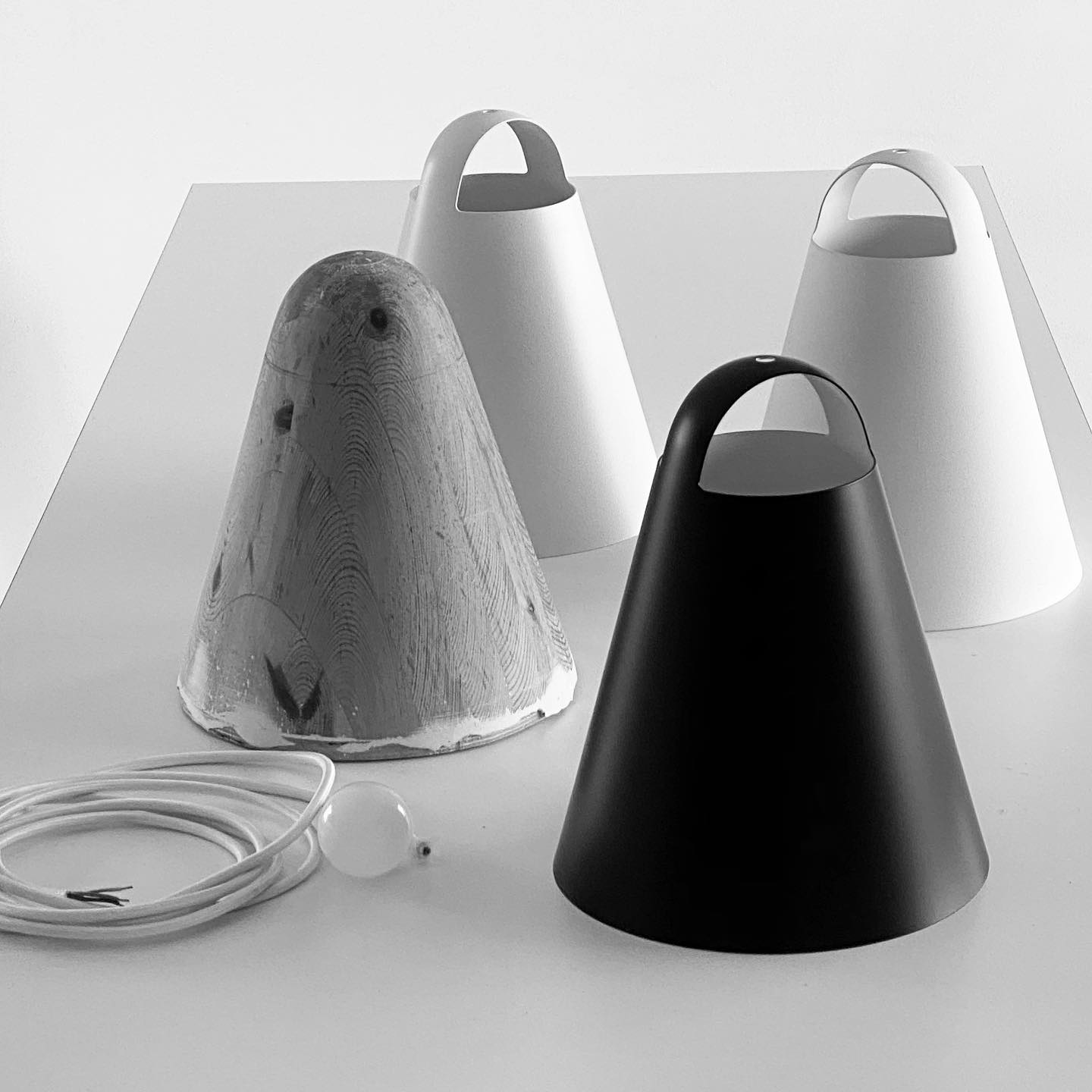 Black and white overhead view of several 'Above' lampshades. Four lampshades are arranged on a light-colored table. One is a prototype and made out of unpainted wood. The other three are final products, two are white and one is black. The shapes are conical, with a slightly wider base and a narrower top. Except for the wooden prototype, each has a small handle-like opening at the top of the cone. 