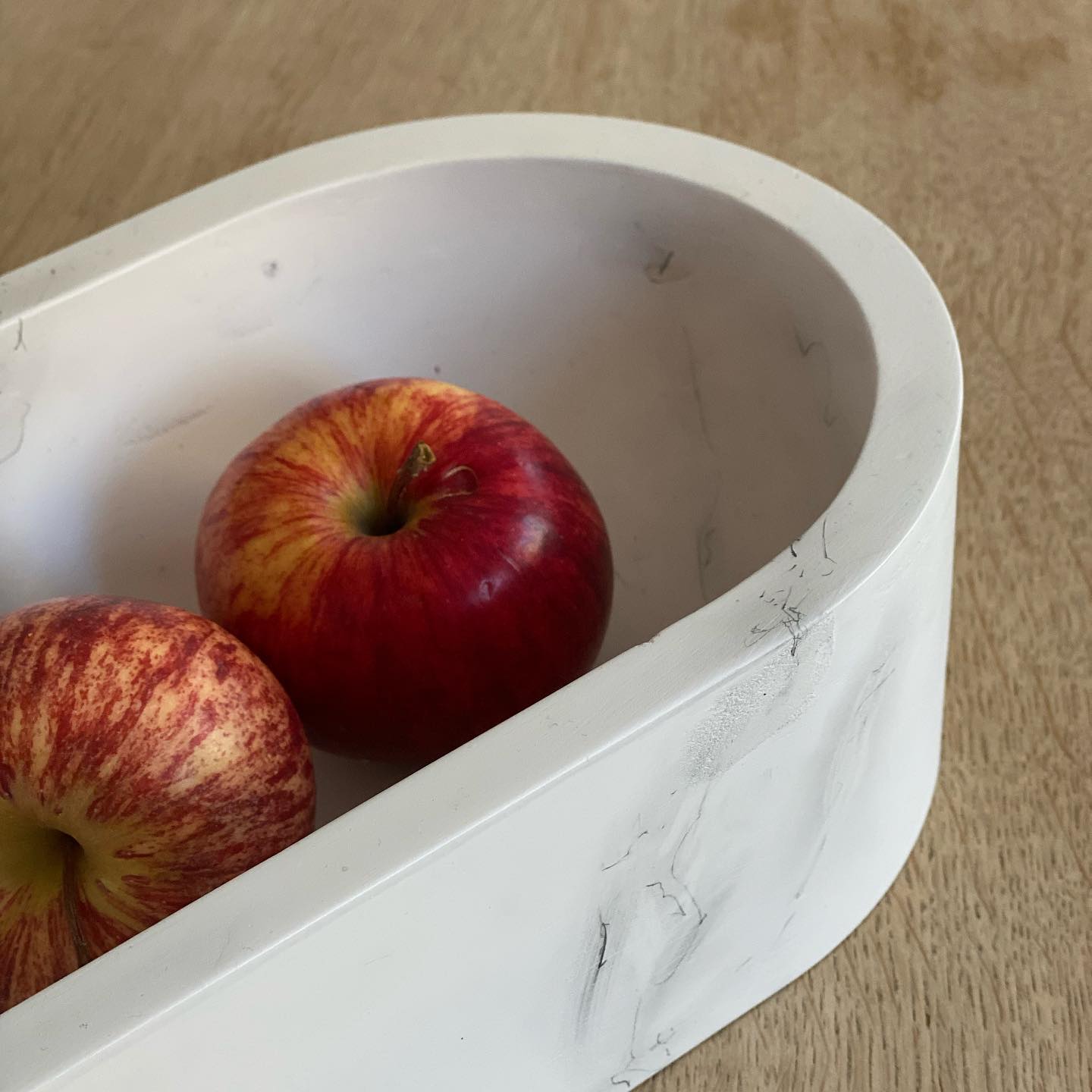 High-angle close-up of a white, marble-patterned bowl with two apples inside. The bowl is a shallow, curved, oblong shape, resembling a half-ellipse. The bowl has thick walls. One apple is a deep red with stripes of lighter red. The other apple is a similar reddish-pink colour, with a more concentrated and less striped pattern of lighter red and pinkish tones. Both apples appear to be fresh and ripe. The background is a light wooden table.