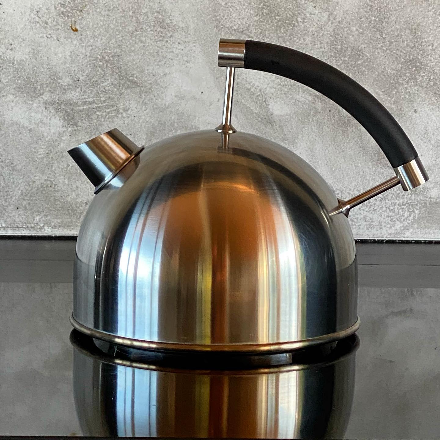 Full shot of a stainless steel kettle. The kettle is round and bulbous, with a smooth, polished stainless steel finish. It has a dark-colored, likely black, curved handle that extends from the top of the kettle to a spout at the back. The spout is also made of the same stainless steel as the body. A small, angled spout protrudes from the top front of the kettle. The kettle sits on a dark surface that reflects the kettle's image. The background is a speckled, light gray concrete-like surface with a slightly rough texture. 