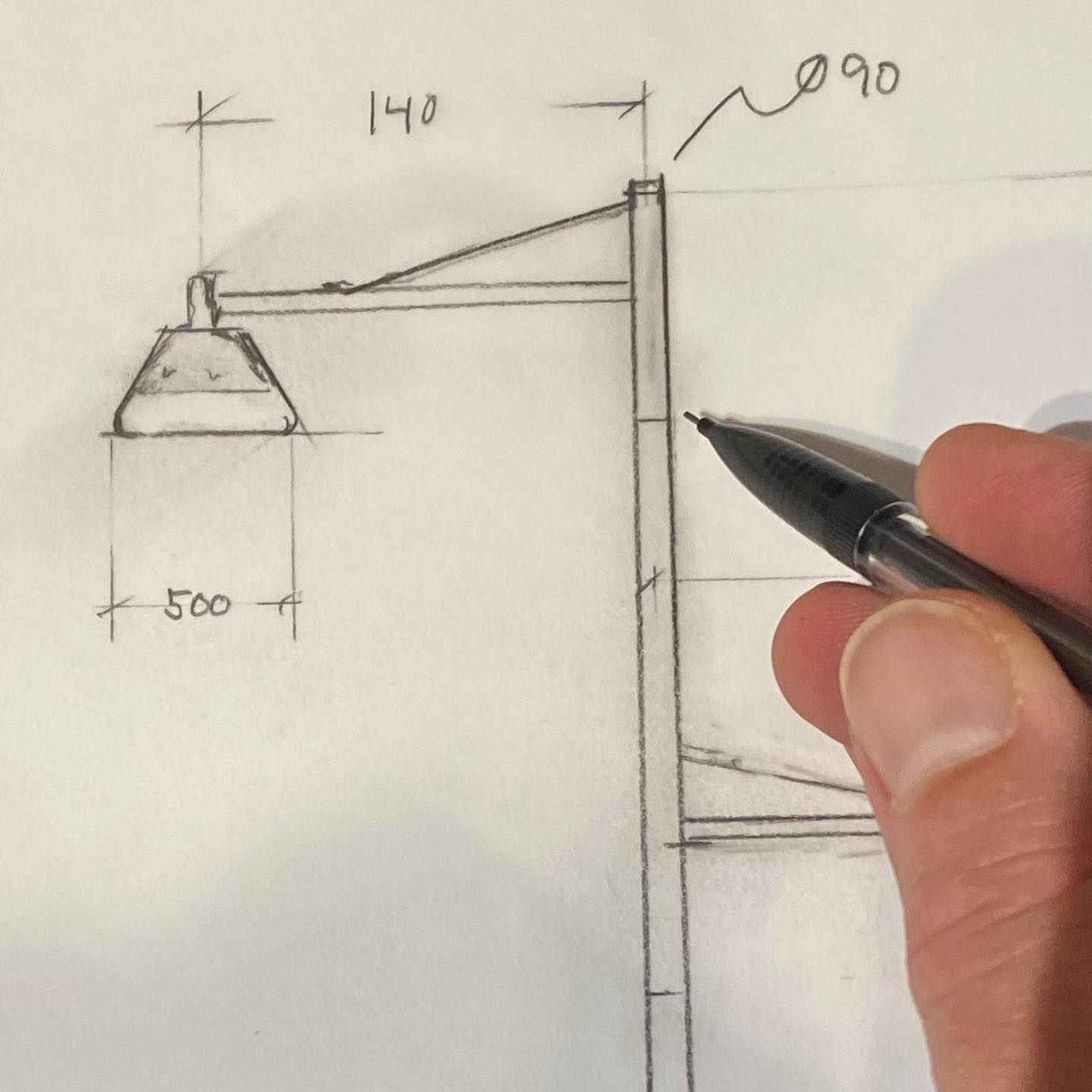 Close-up of a pencil sketch on paper. The sketch is a design plan for a street lamp. The street lamp is drawn with a conical or pyramid-shaped shade. Dimensions, marked as '140' and '500', are indicated with reference lines. Mads Odgård's hand can be seen holding a black pen, seemingly used to add further annotations or fine-tune the drawing.