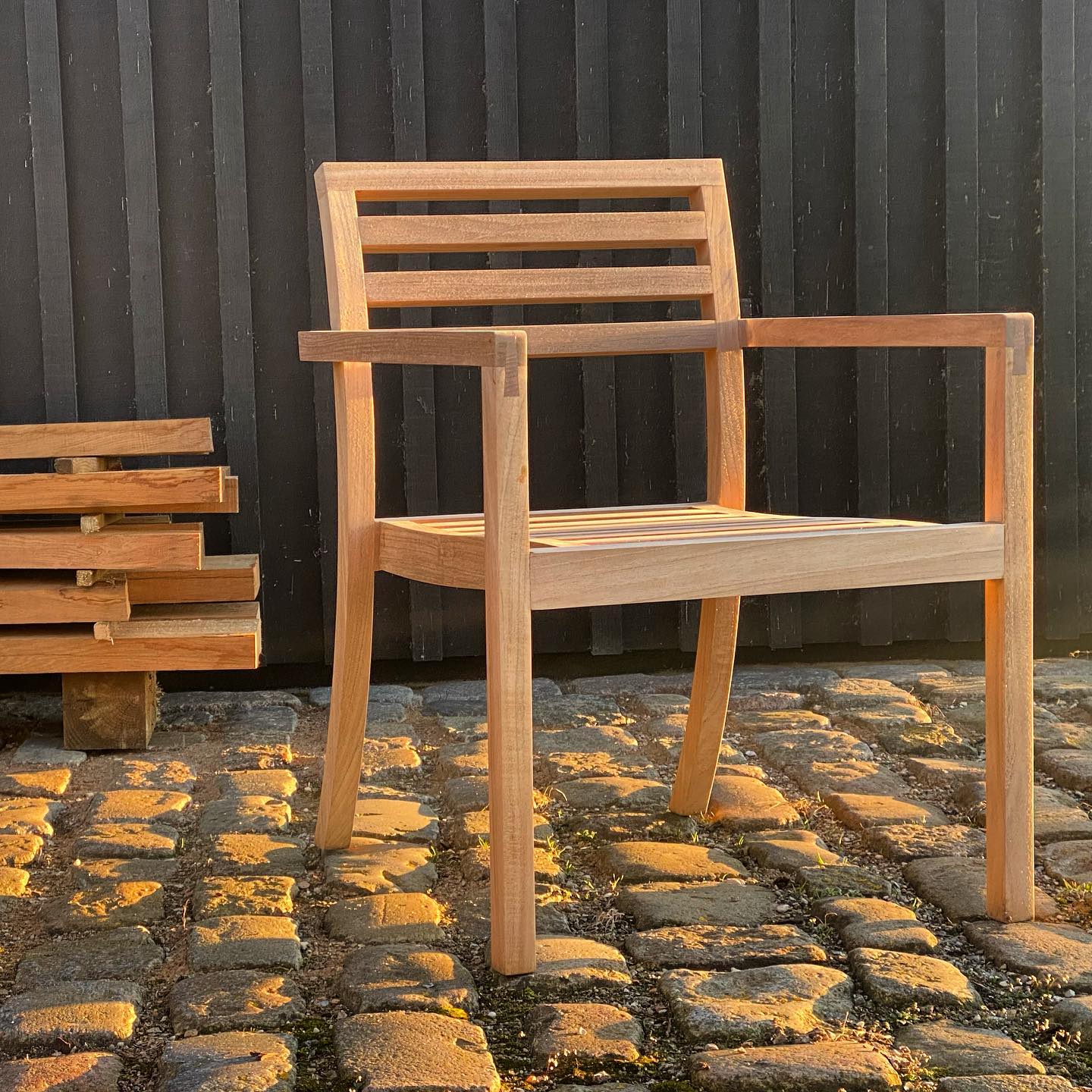 Full outdoor shot of a light-brown wooden chair sitting on a cobblestone surface. The chair is a simple design with a slatted back and straight lines.  Its legs are straight and substantial, made of the same wood as the rest of the chair. The chair's seat and back are constructed from slats arranged in a grid pattern. The cobblestone ground is illuminated by the sun, giving a textured look to the paving. Some small patches of green vegetation or weeds are visible growing between the stones. In the foreground, to the left of the chair, a simple wooden bench or low table made of stacked, horizontal planks is visible. A dark, vertical-plank wooden wall stands behind the chair, acting as a backdrop.A stack of planks lay infron of the wall to the left of the chair.