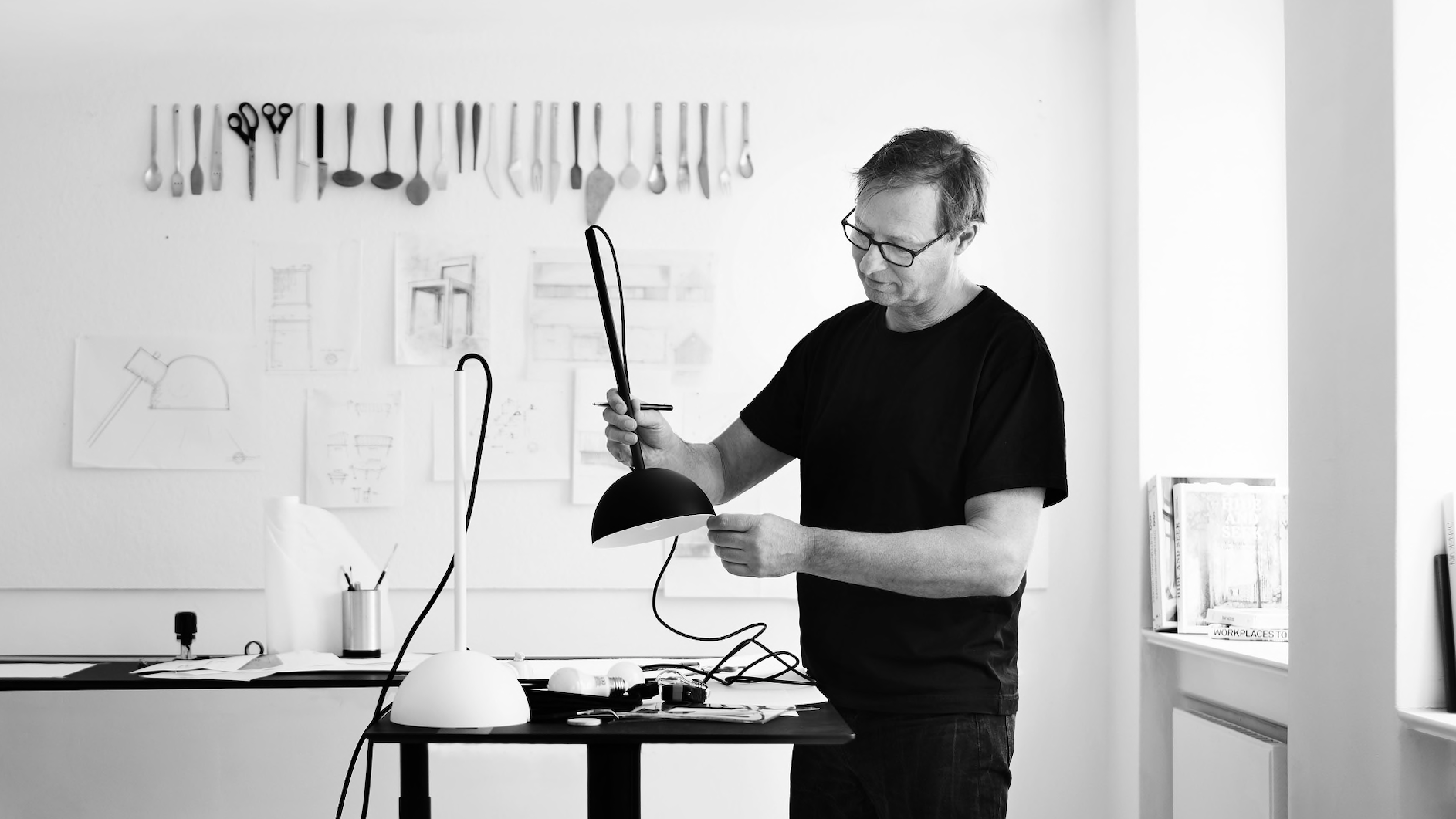 Full black and white indoor shot of Mads Odgård working at a desk in a design studio. He is slightly angled toward the lamp, wearing a black, t-shirt. He is wearing glasses, looking downward, his attention directed at the lamp he is working with. Mads' hands are visible, engaging with the lamp. His posture suggests attentiveness and focus on a task. The workspace is a stark, minimalist design studio.  A white wall is hung with a collection of various utensils (knives, forks, spoons, and scissors) displayed on the wall. Beneath the wall-mounted utensils, there are sketches and design plans on the wall in the form of rough, hand-drawn diagrams and architectural renderings. 