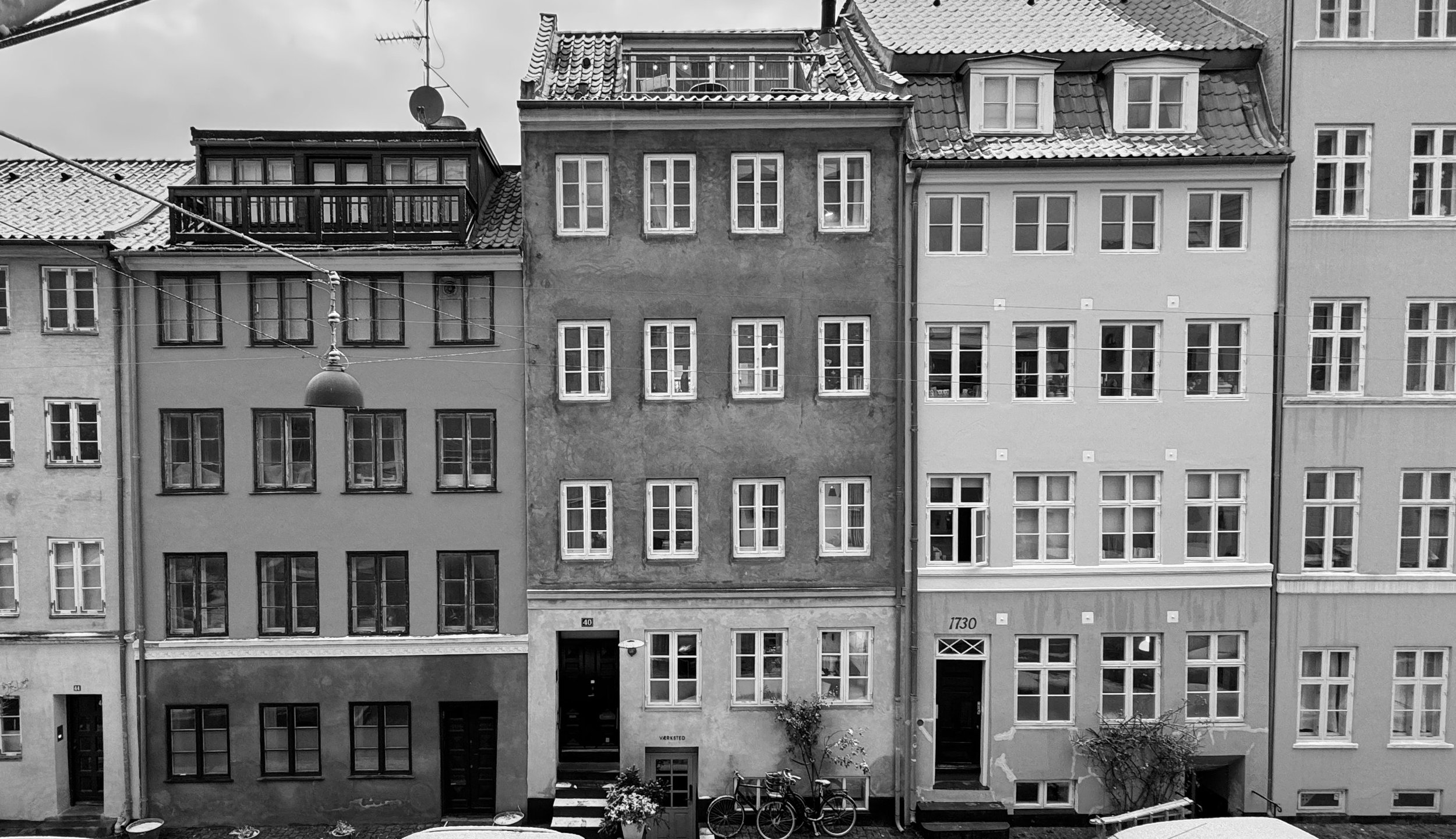 Full black and white shot of a row of buildings. Several multi-story buildings are aligned closely together. Mads Odgård's design studio is the house in the center of the picture. Each building has a series of evenly spaced windows along its facade, creating a regular pattern. The windows are rectangular and appear to be of similar size and style throughout. The buildings' exteriors are composed of a light neutral tone. A street-level lamppost is visible near the center of the image.